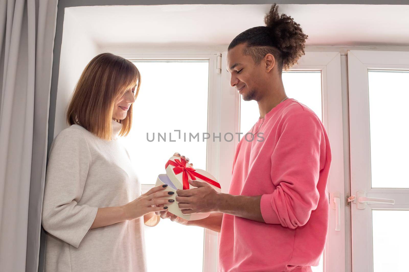 A happy guy in a pink jumper and a girl in a white dress are standing at a large white window. The guy presented a surprise - a white box in the shape of a heart with a bow. close up