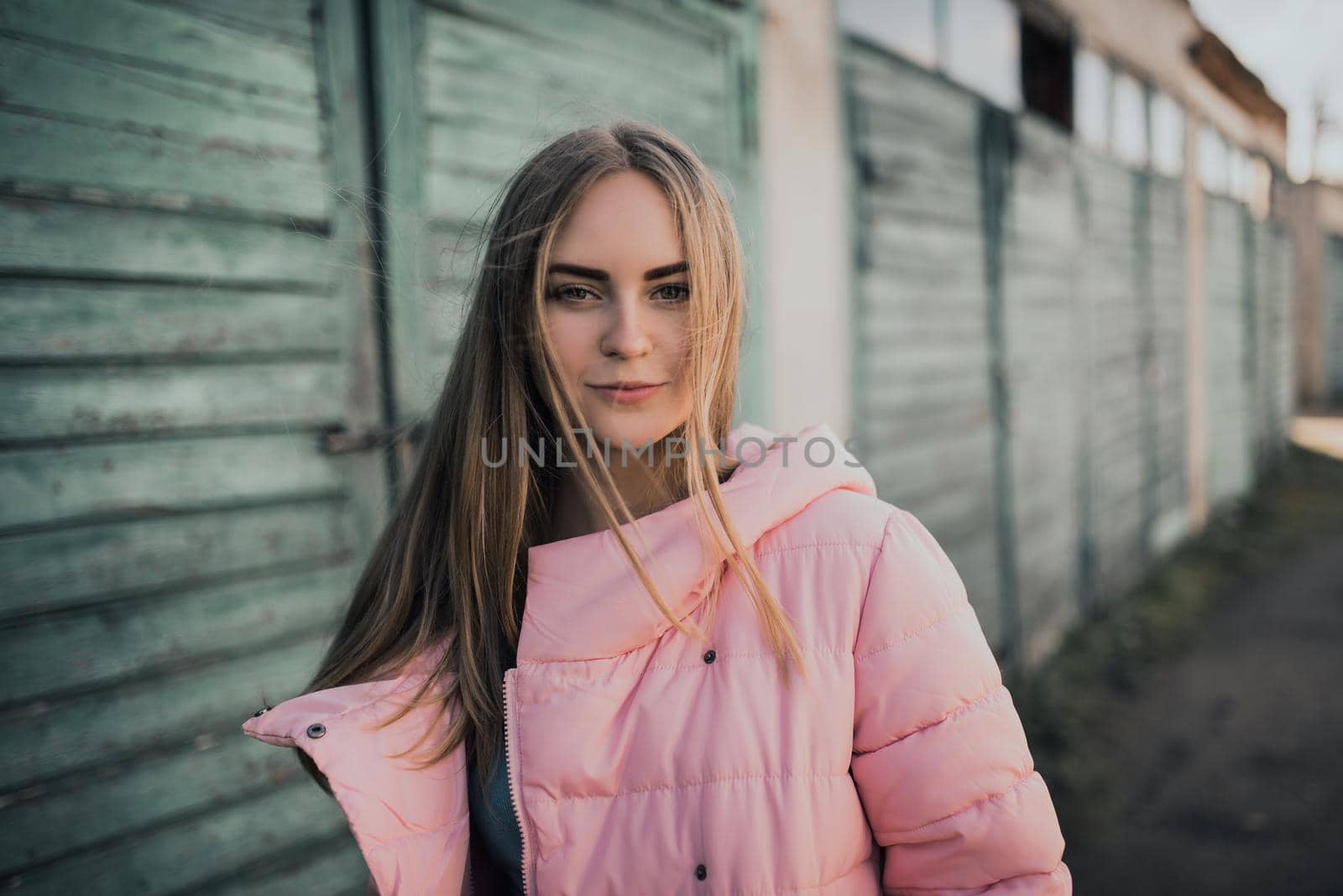 Young gorgeous blonde girl dressed fashion pink jacket and blue jeans. Old azure cerulean fence on the background. Picture ideal for illustrating women's magazines.