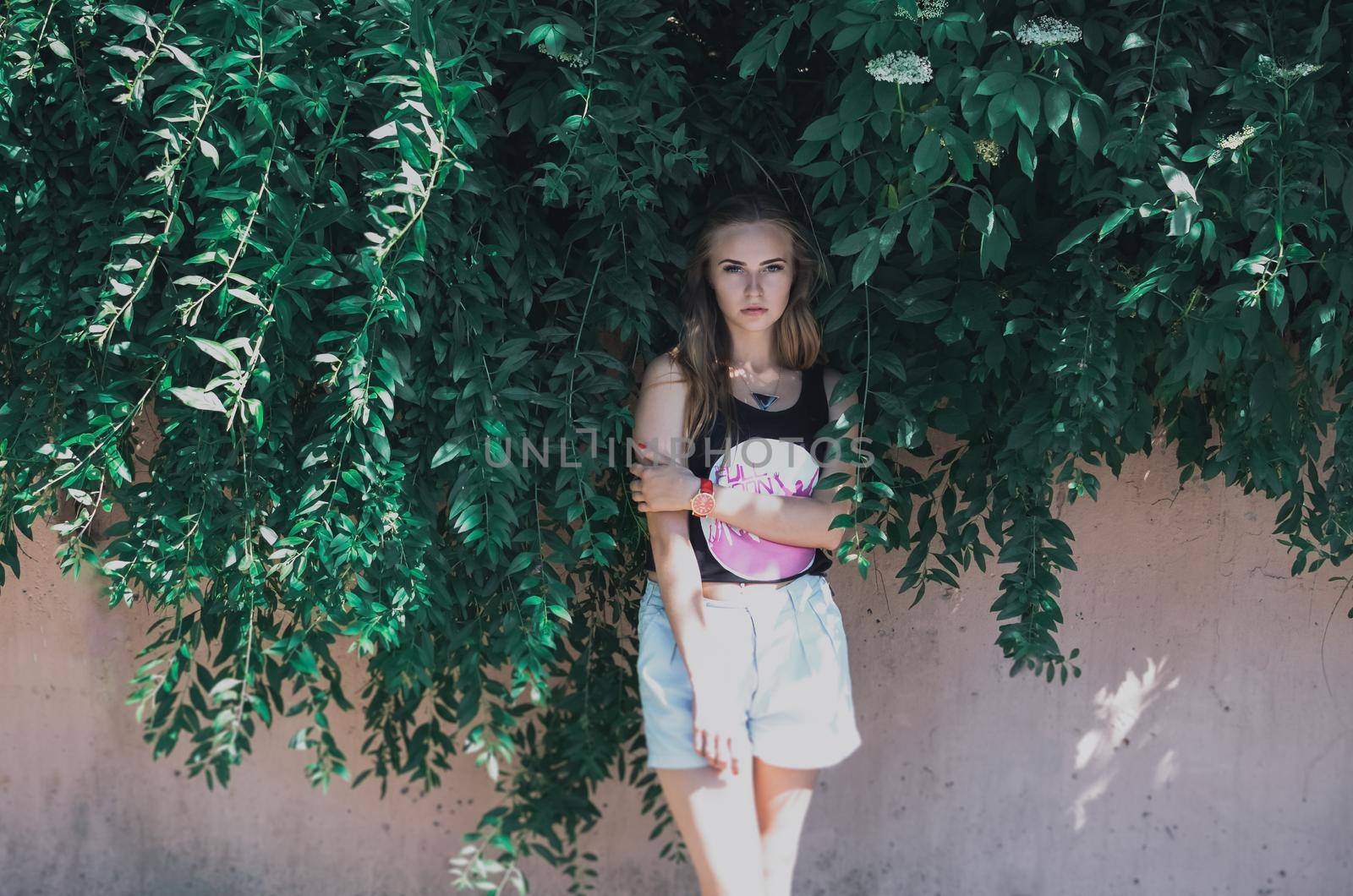 Lovely young blonde girl with long curly hair on a background of green thick leaves. A woman is wearing a cropped black top with a white print and pink lettering and high-waisted shorts.closed eyes