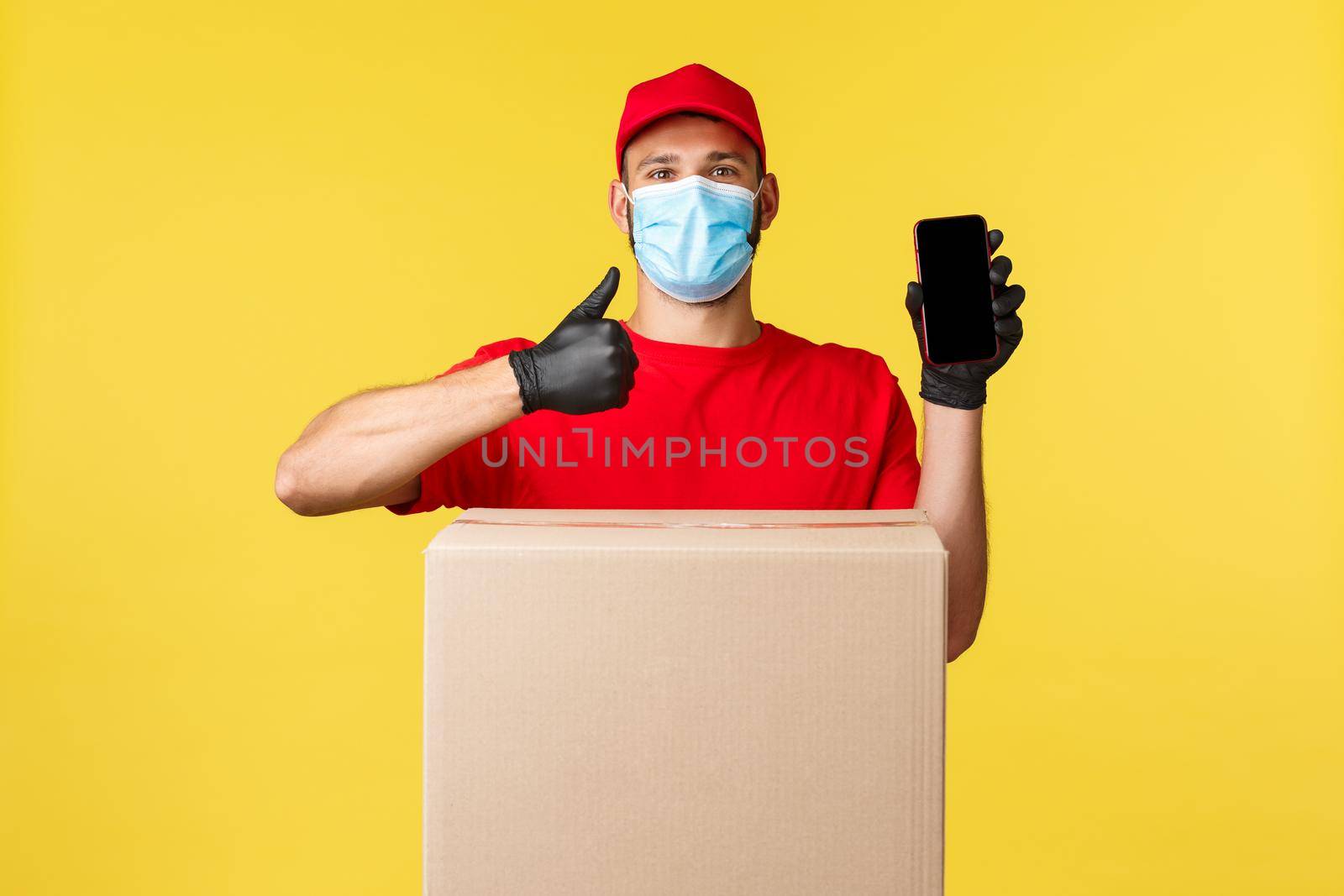 Express delivery during pandemic, covid-19, safe shipping, shopping concept. Courier in red uniform, showing deliver service app, thumb-up, stand near box package, application for tracking order.