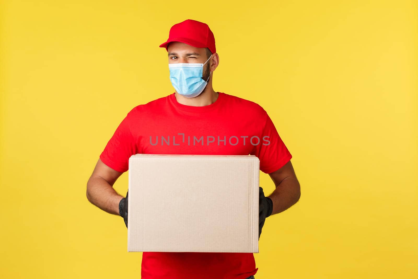 Express delivery during pandemic, covid-19, safe shipping, online shopping concept. Cheeky smiling courier in red uniform and medical mask, wink, holding parcel box, deliver order.