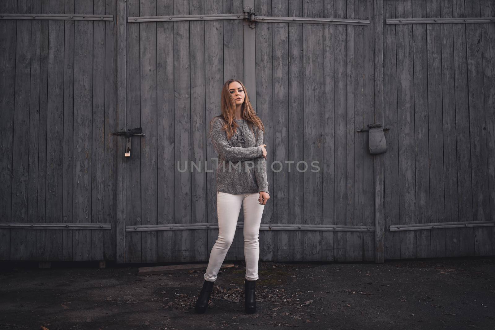 beautiful young blonde model girl. White pants. gray knitted sweater. black boots. wooden pendant on the neck in the form of a horse. On the Sunset. on a background of gray wooden gates