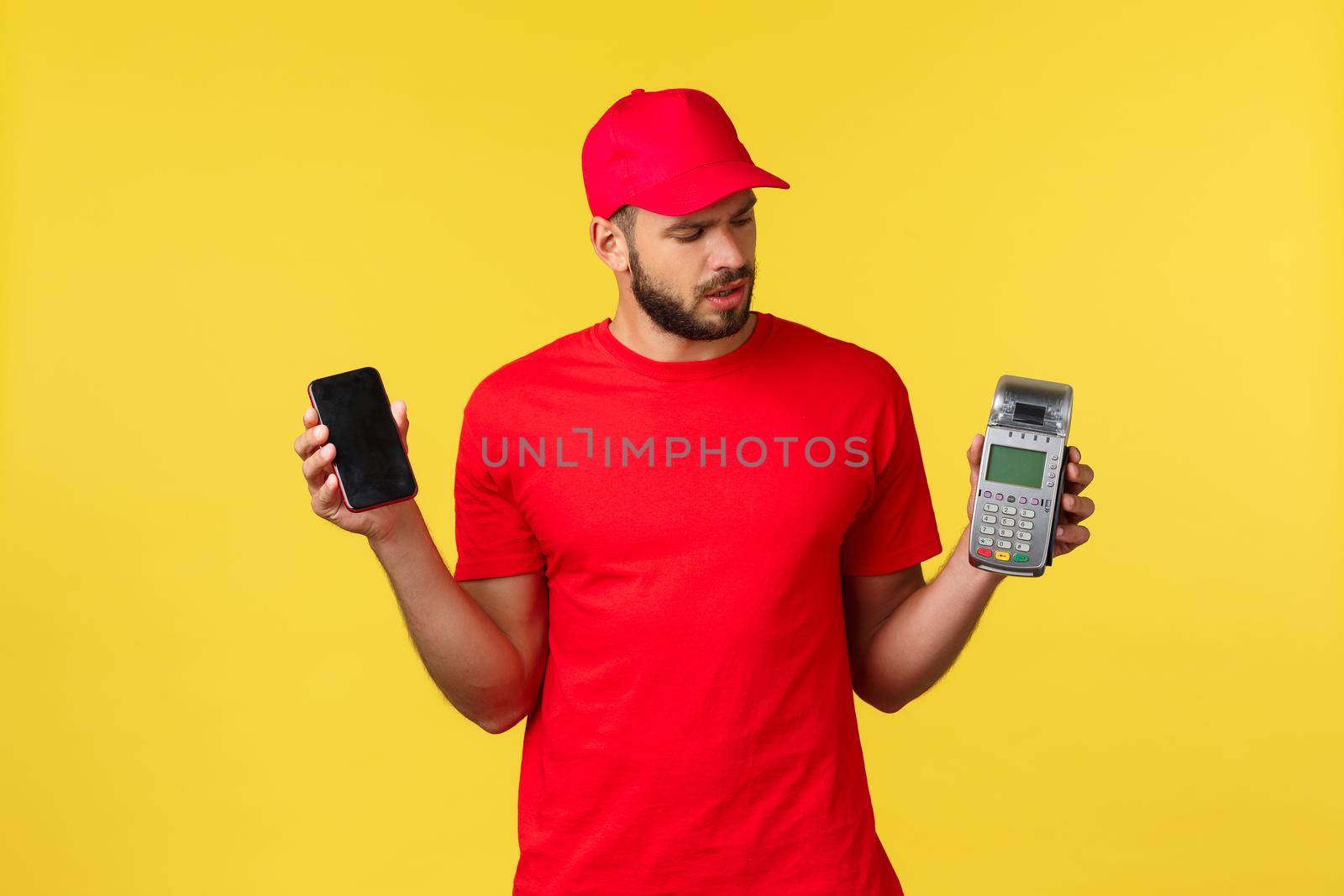 Contactless payment, online delivery, internet shopping concept. Confused young courier in red uniform, newbie courier figure out how POS terminal works, holding mobile phone.