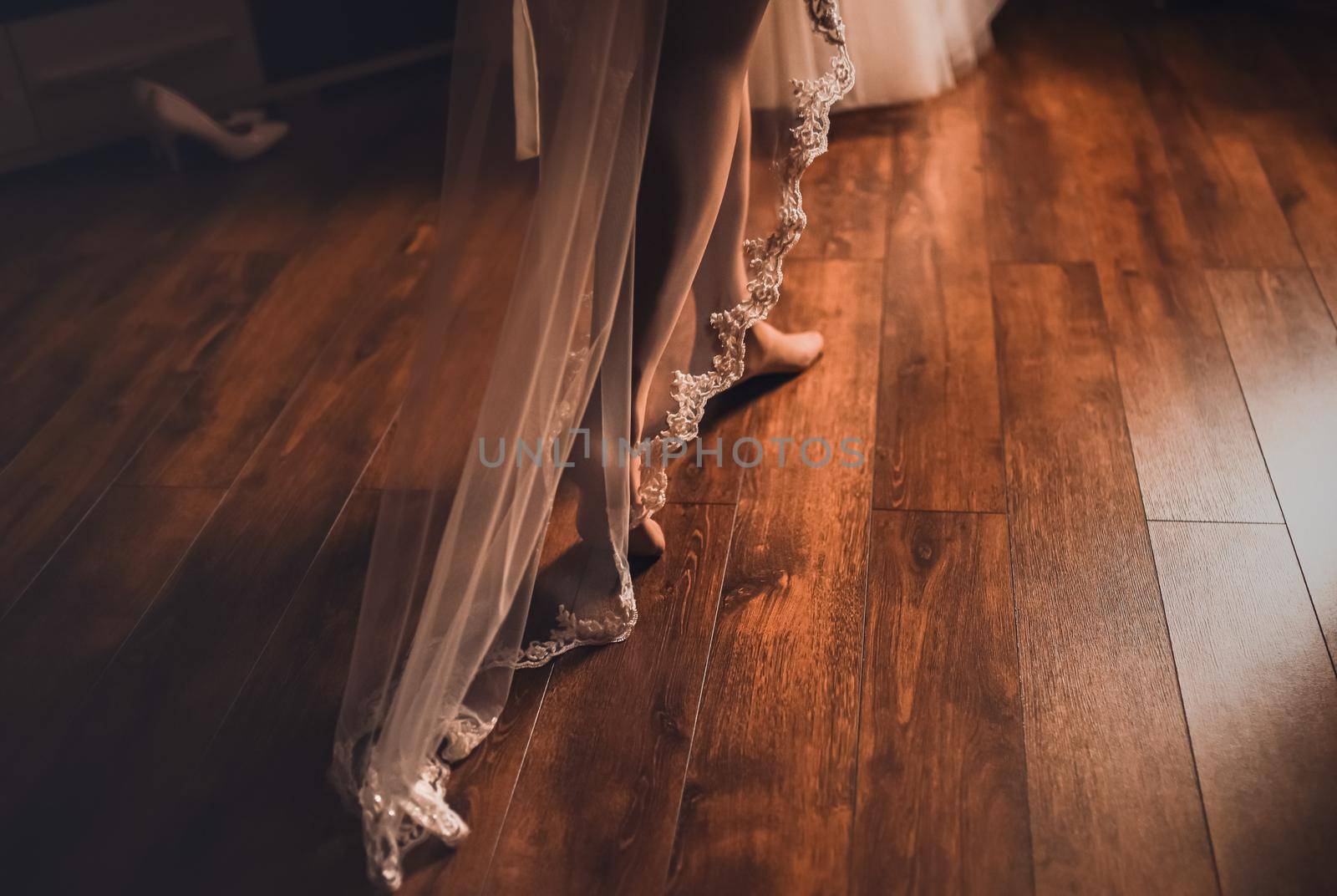 The bride's legs in white stockings walk along the brown wooden parquet floor, there is a long lace veil in the back. dark background