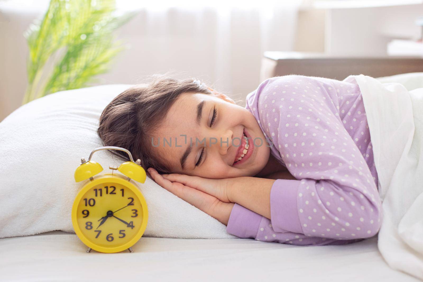 A adorable smiling brunette girl in purple polka dot pajamas lie on a white pillow, on a bed, in the morning light, a yellow alarm clock stands nearby. copy space