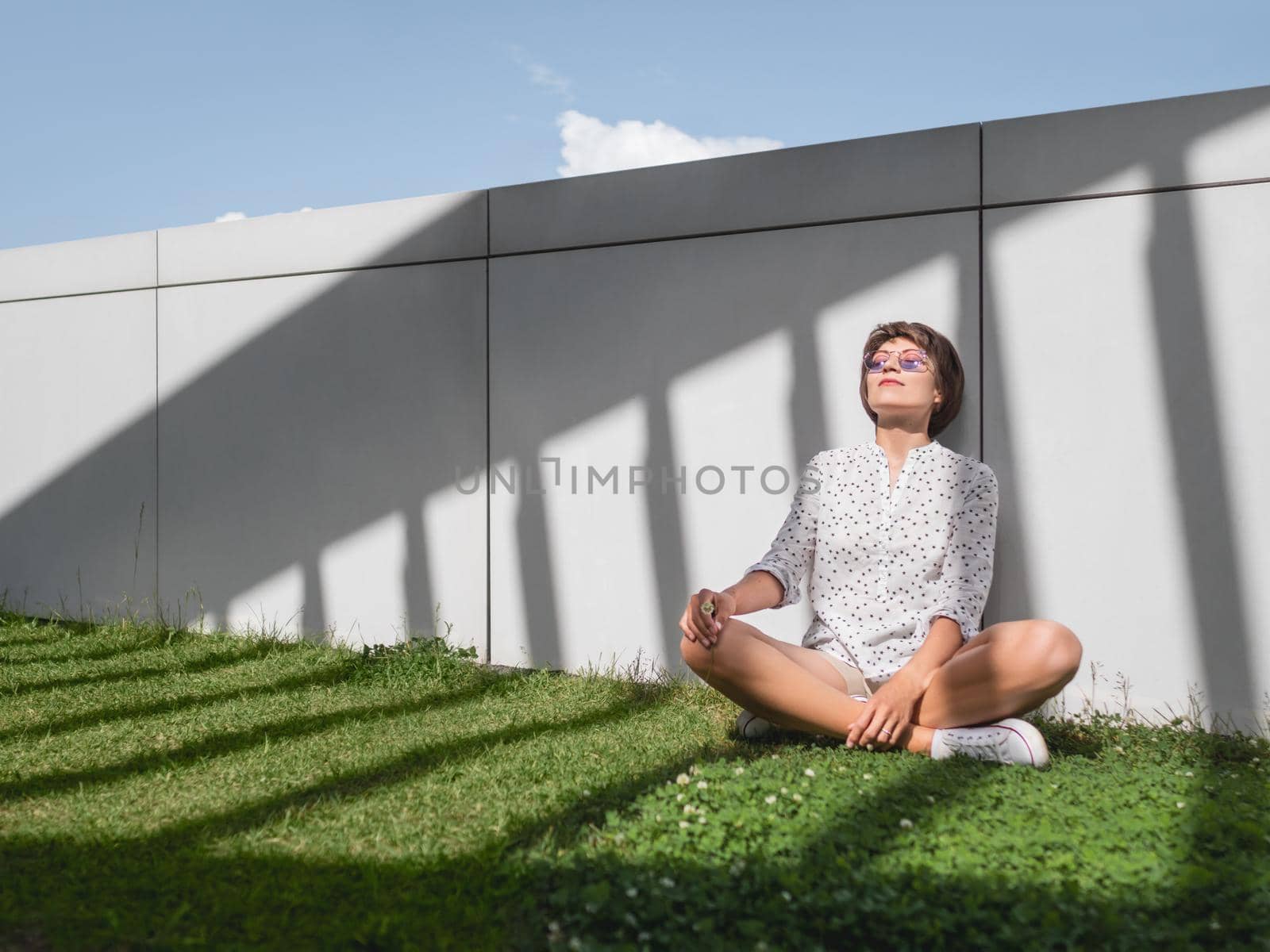 Woman is meditating on lawn in urban park. Nature in town. Relax outdoors after work. Summer vibes.