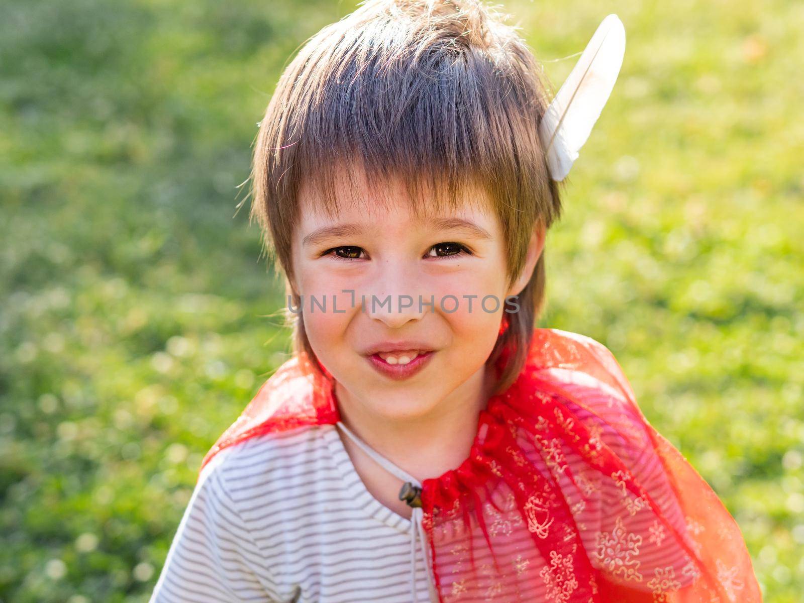 Portrait of smiling boy playing American Indian. Kid with white bird feather and red cloak. Costume role play. Outdoor leisure activity.