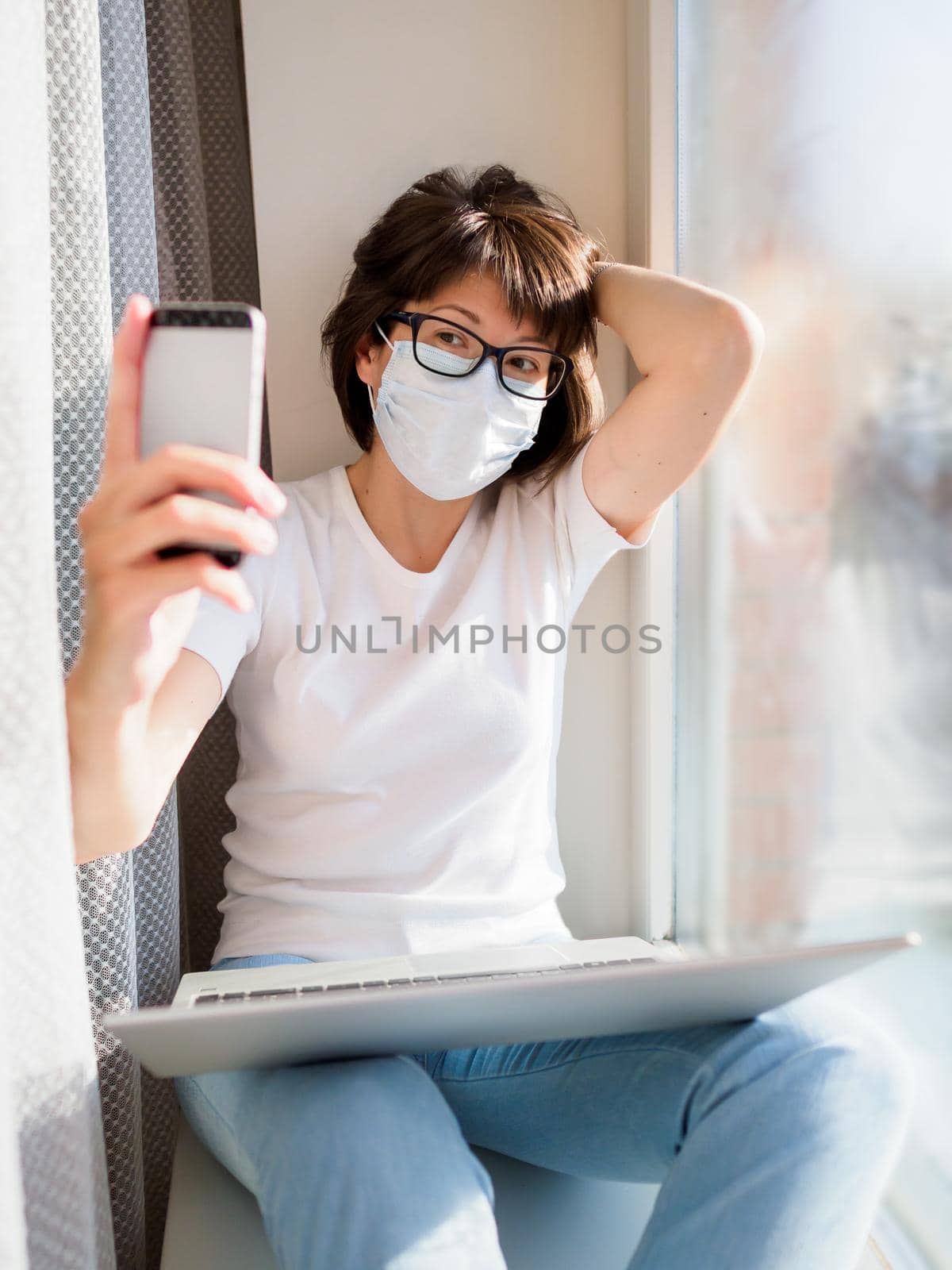 Pretty woman in medical mask remote works from home. She is making selfie on window sill with laptop on knees. Lockdown quarantine because of coronavirus COVID19. Self isolation at home.
