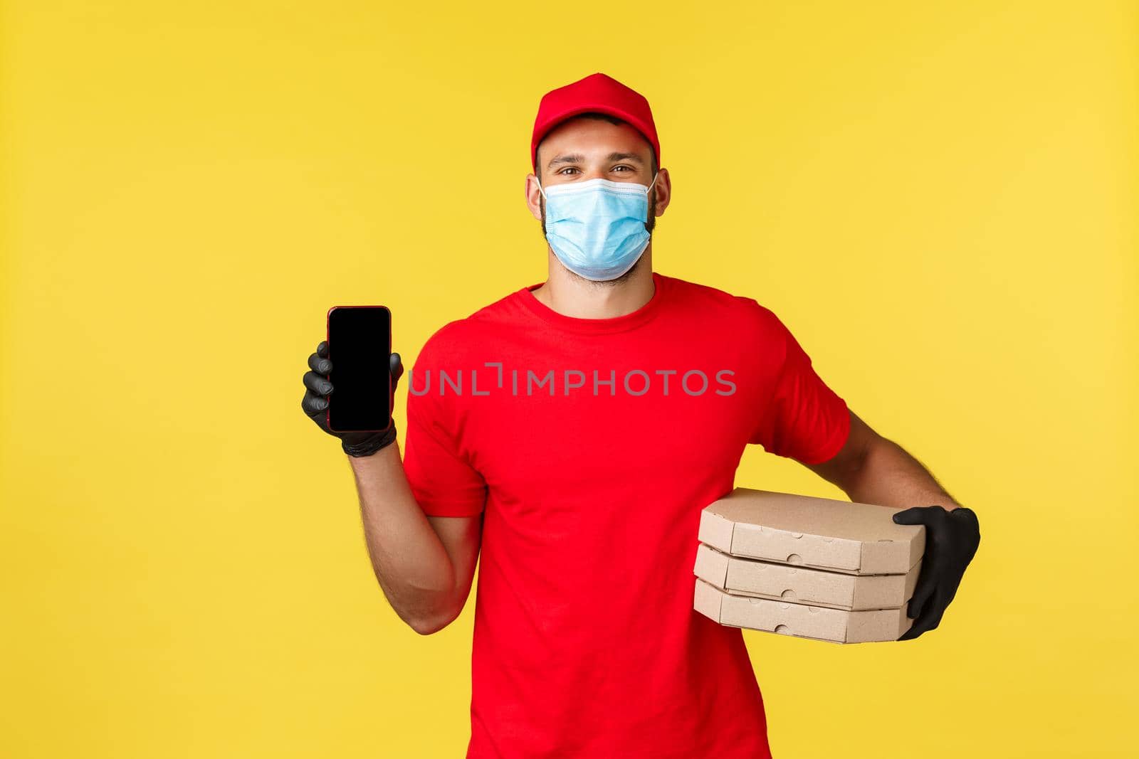Food delivery, tracking orders, covid-19 and self-quarantine concept. Friendly smiling courier in red uniform, medical mask and gloves, showing smartphone screen application, bring pizza.