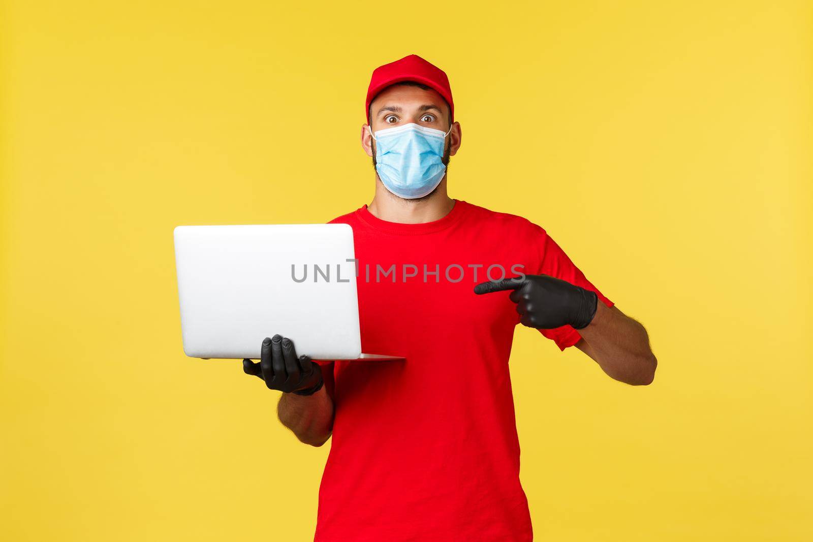 covid-19, safe shopping, express delivery and order tracking concept. Surprised courier or employee in red uniform, medical mask, pointing finger at laptop with impressed excited face.