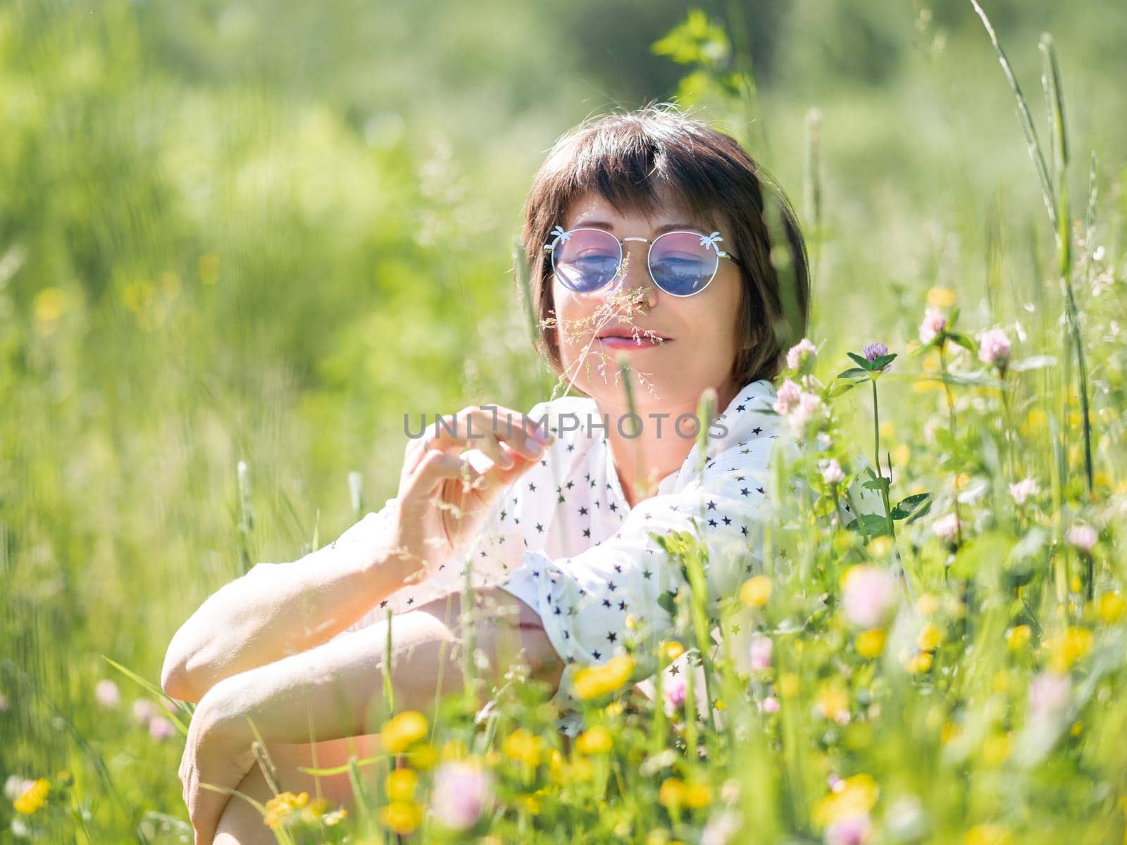 Woman in colorful sunglasses, enjoys sunlight and flower fragrance on grass field. Summer vibes. Relax outdoors. Self-soothing. by aksenovko
