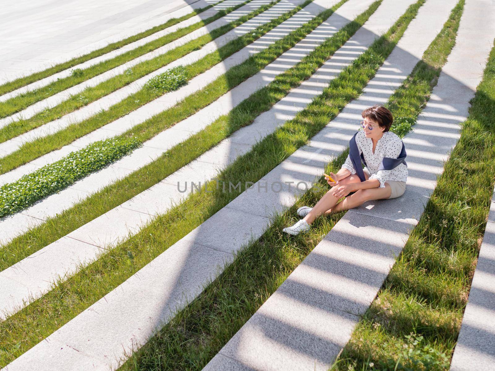 Woman texting in her smartphone while sits on bench in urban park. Striped shadow on green lawn. Modern lifestyle of millennials.