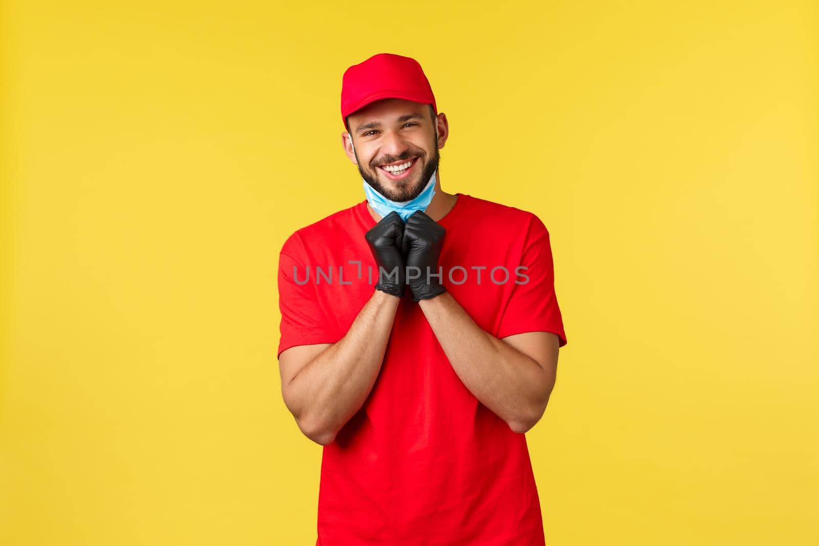 Express delivery during pandemic, covid-19, safe shipping, online shopping concept. Friendly charismatic courier in red uniform, gloves, take-off medical mask to smile sincere, yellow background.