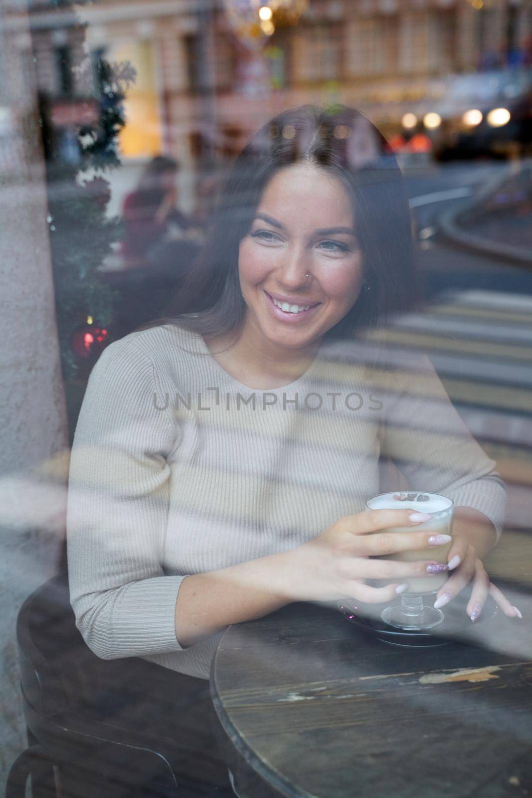A beautiful girl sits in a cafe and looks out window thoughtfully. Reflection of city in window. Smiling brunette woman with long hair drinks cappuccino coffee, vertical by NataBene