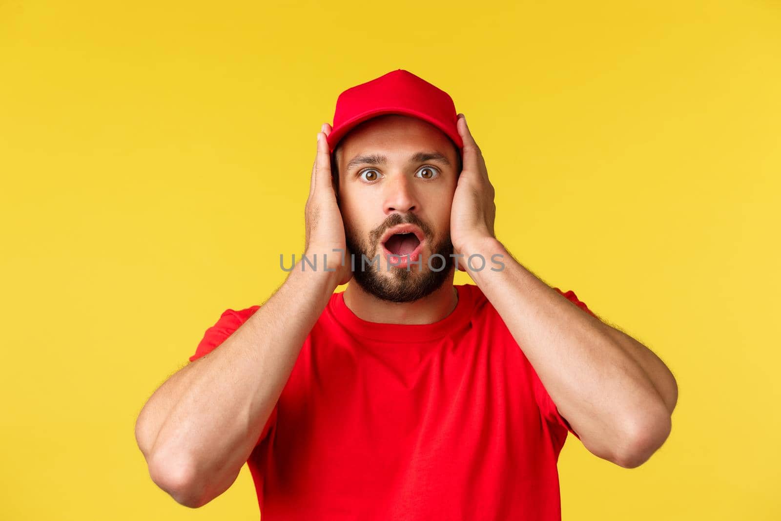 Close-up of shocked gasping male courier, employee in red uniform cap and t-shirt, cover ears and staring speechless at camera, heard stunning news, standing yellow background.