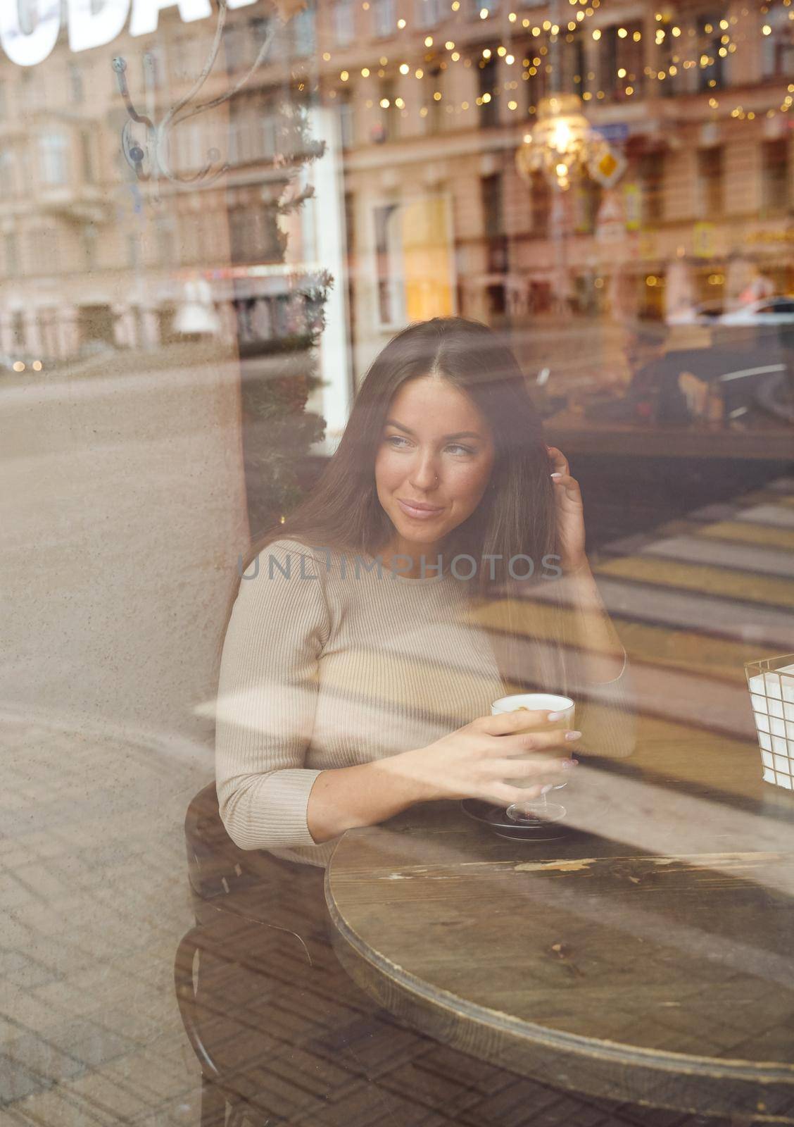 A beautiful girl sits in a cafe and looks out window thoughtfully. Reflection of city in window. Brunette woman with long hair smiling and drinking cappuccino coffee, vertical