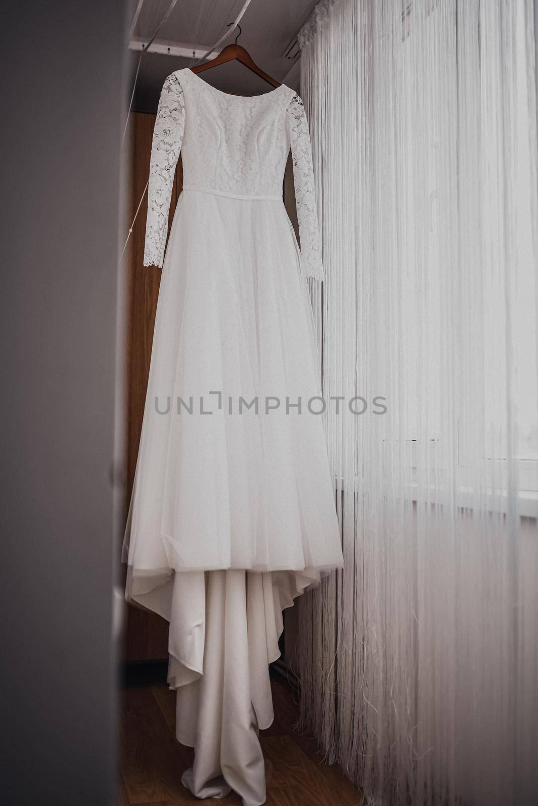 A white luxurious beautiful wedding dress hangs on a hanger. Pleated white chiffon curtains hang around.