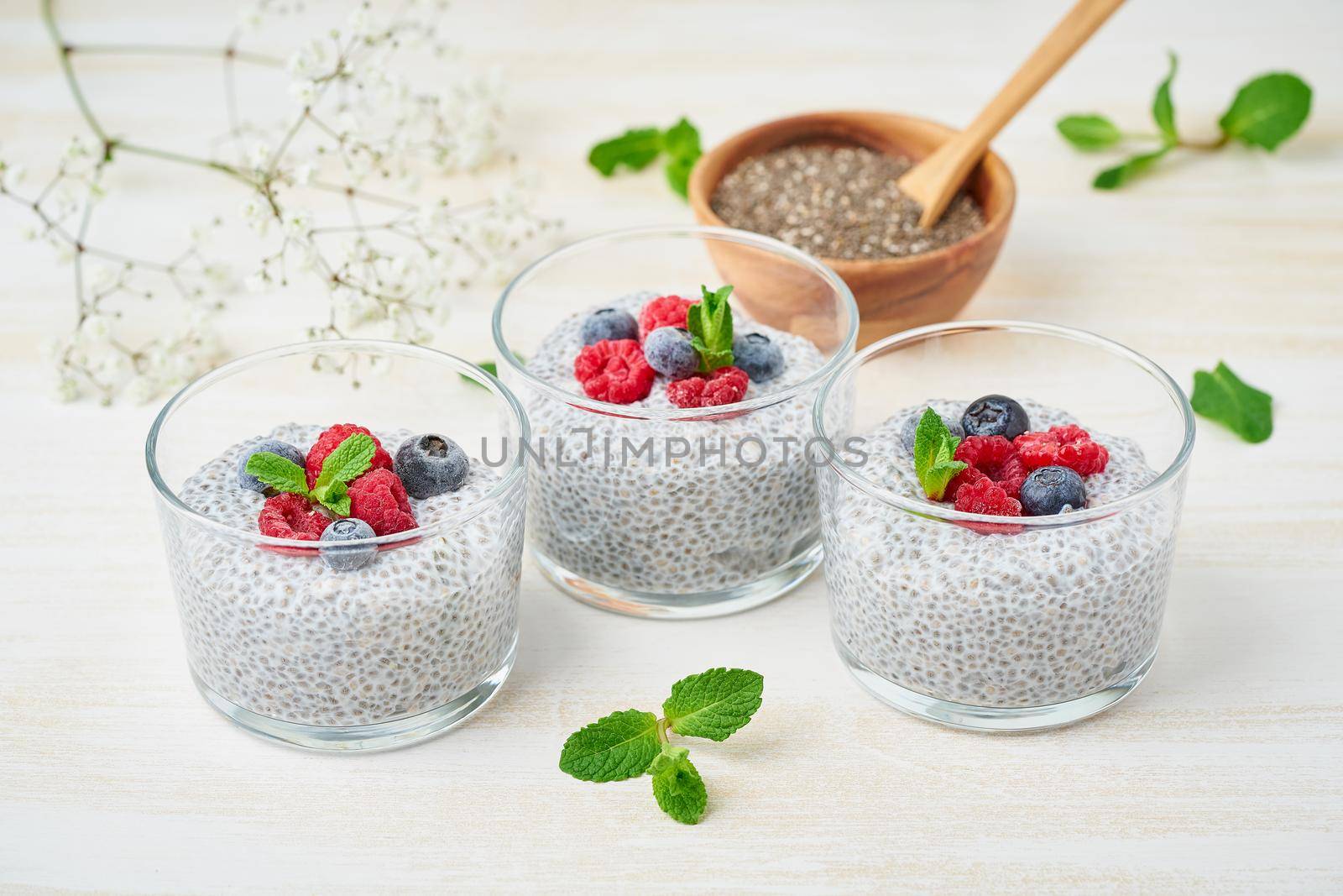 Chia pudding with fresh berries raspberries, blueberries. Three glass, light wooden background, side view, flowers
