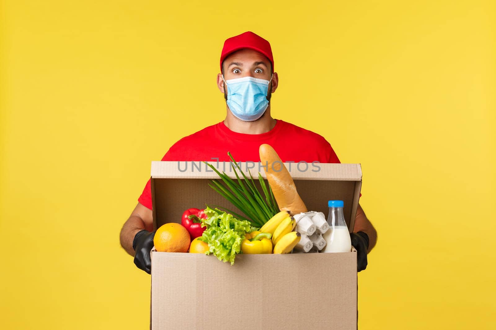 Takeaway delivery, covid-19 quarantine and groceries concept. Surprised courier in medical mask, uniform red cap and t-shirt, staring shocked, bring food order box to customer home, yellow background.