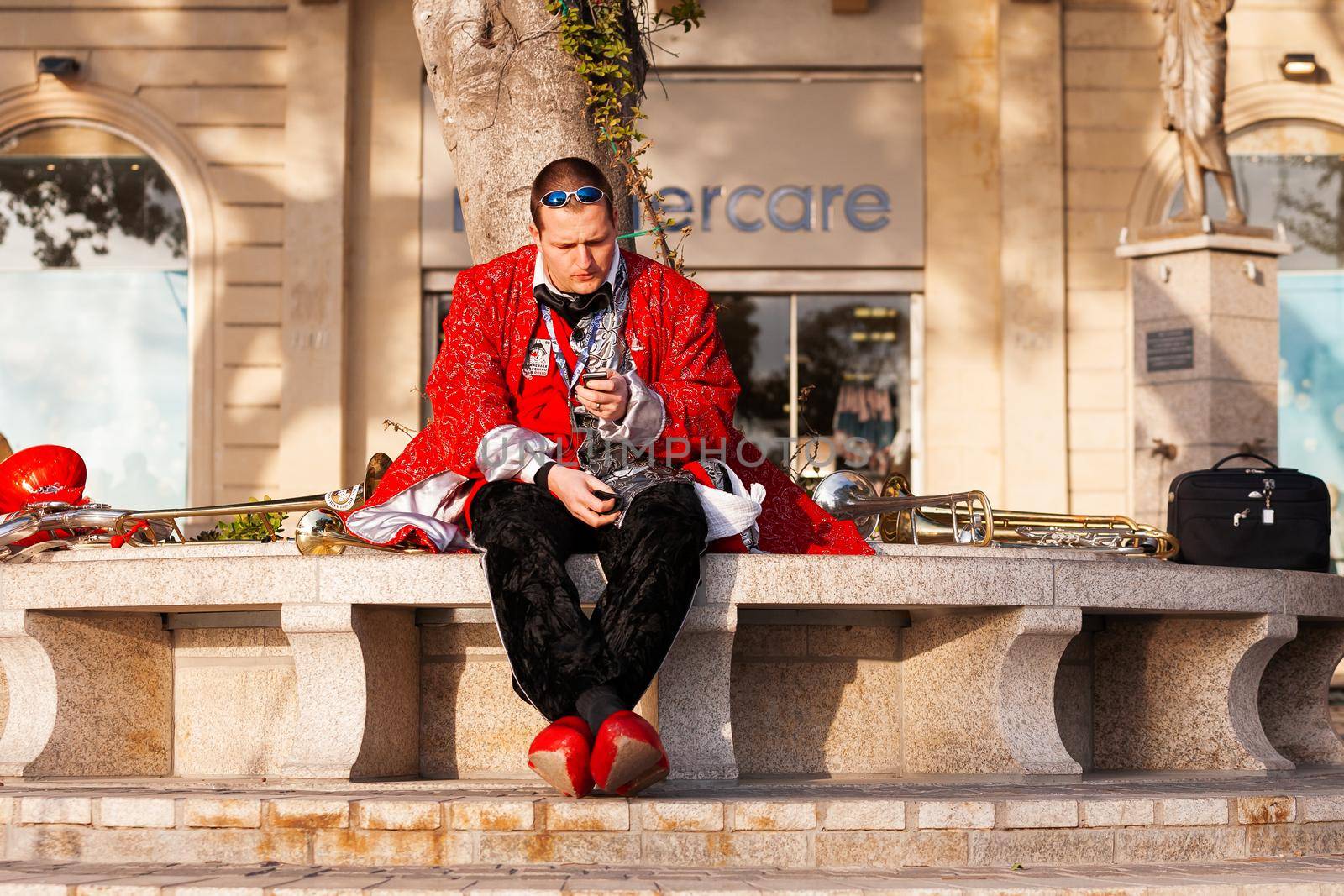 SLIEMA, MALTA - February 13, 2010. Musician with smartphone is sitting on bench. Man in colorful costume is ready for night carnival. by aksenovko