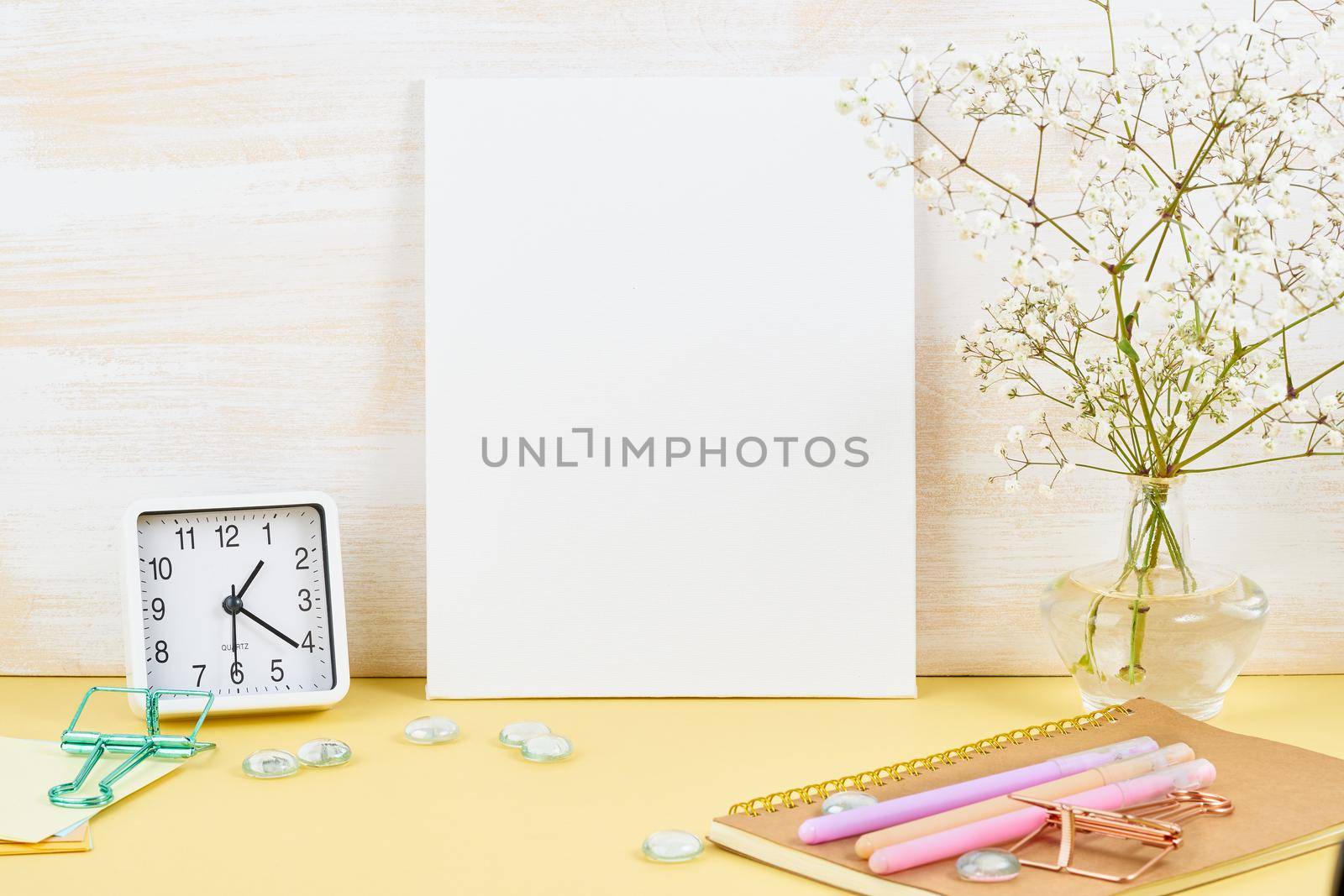 Mockup with blank white frame on yellow table against wooden wall, alarm, flower in vaze.