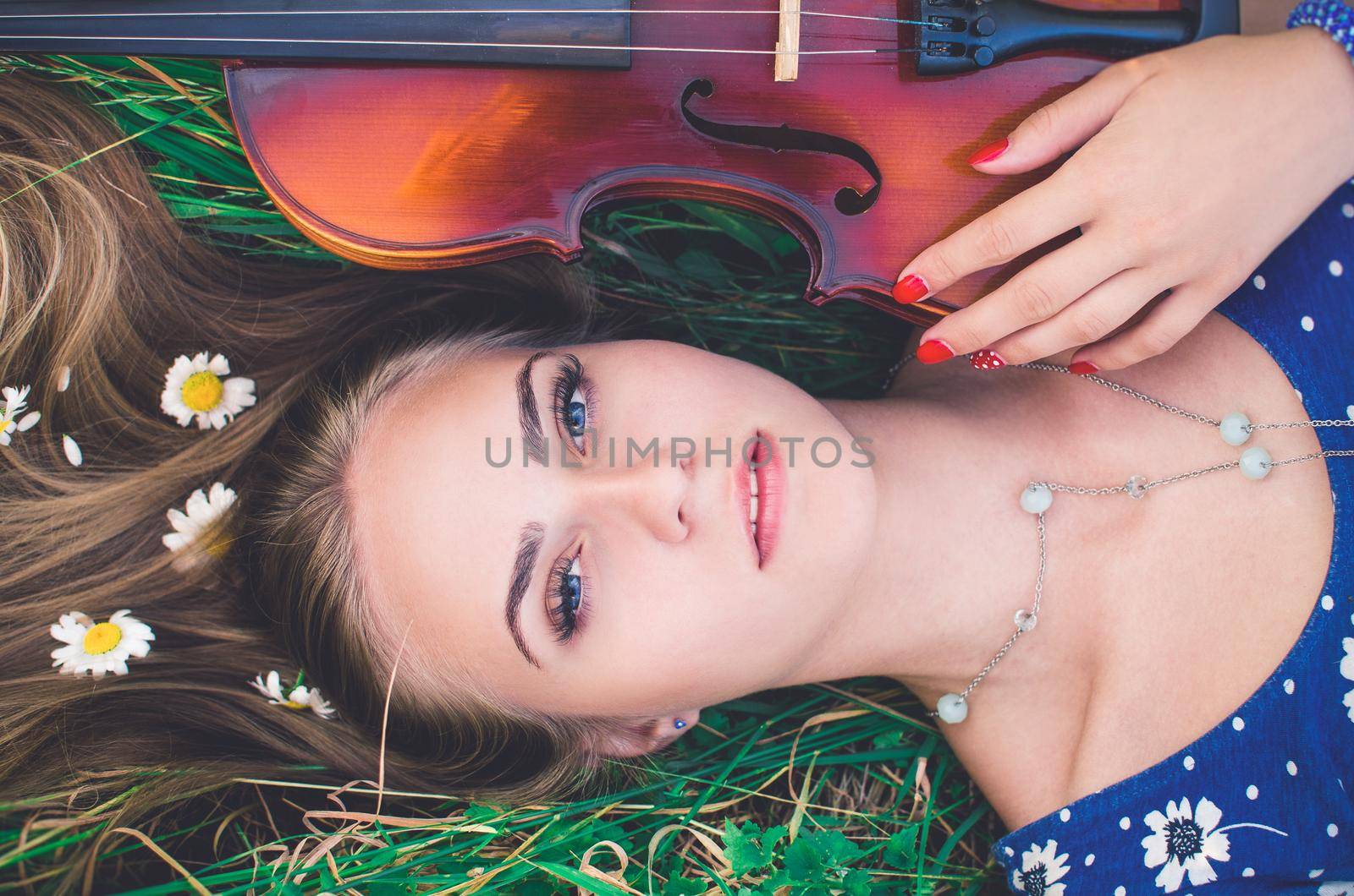 young slim fair-skinned girl blonde beautiful lies in tall grass. Girl in a blue short tight dress with a print of daisies. A bouquet and lonely daisies scattered around.A woman hugs violin. Top view