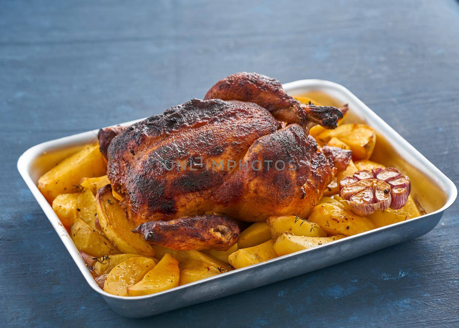 Baked whole chicken in blue casserole on dark blue table, roasted meat with potatoes. Side view.