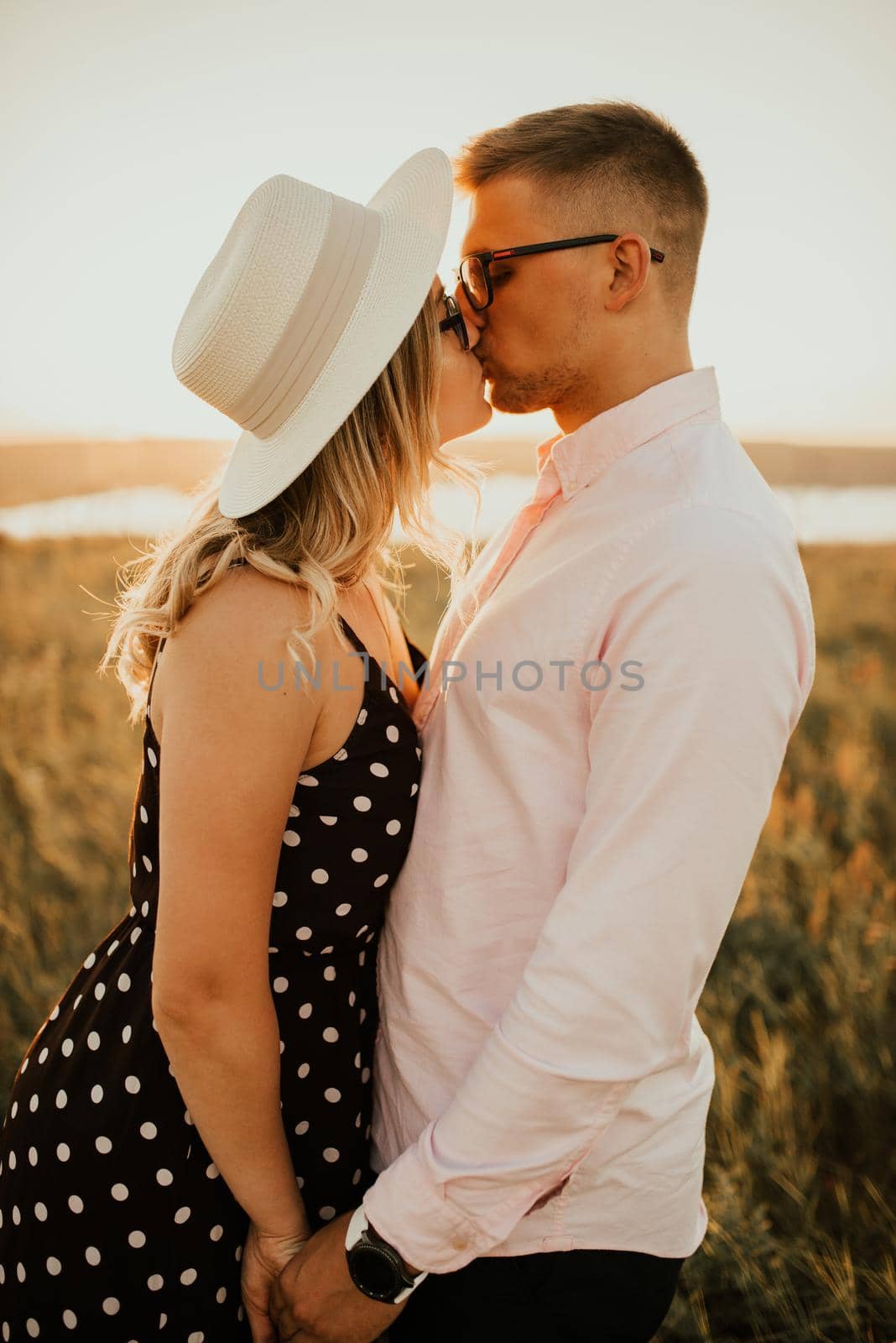 A man with a woman in a hat hug and kiss in the tall grass in the meadow. A couple of fair-haired fair-skinned people in love are resting in nature in a field at sunset.