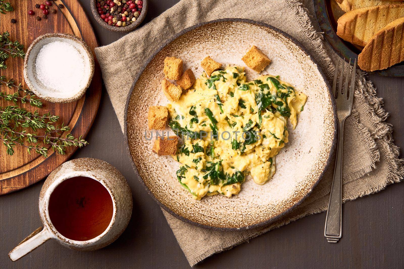 Pan-fried scrambled eggs and spinach, cup of tea on dark brown background. Breakfast with omelette, top view