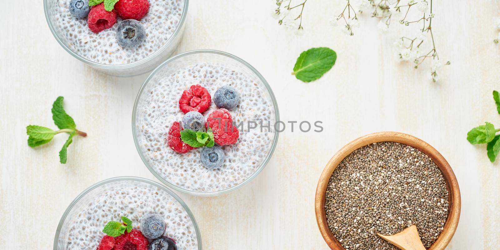 Long banner with chia pudding, top view, fresh berries raspberries, blueberries. Three glass, light wooden background, flowers