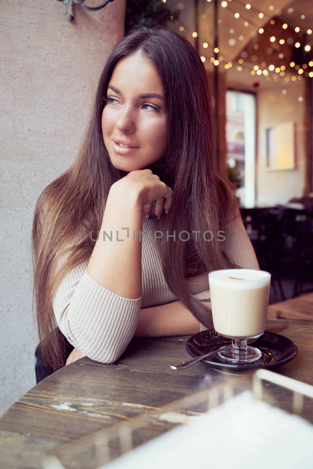 Beautiful girl sits in cafe in Christmas holidays, in background lights of garland. Brunette woman with long hair drinks cappuccino coffee, latte and looks out window, dark backdrop by NataBene