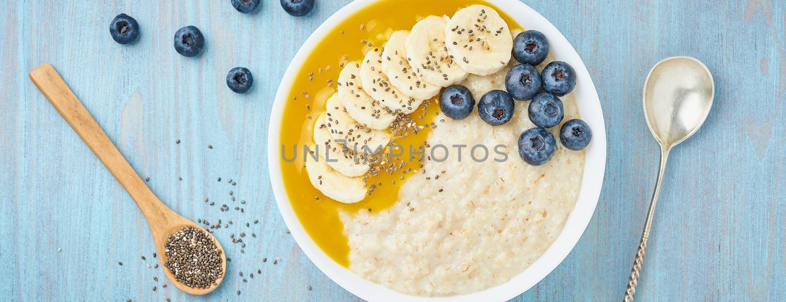 Banner with oatmeal, bananas, blueberries, chia seeds, mango jam on blue wooden background. Top view. Healthy breakfast