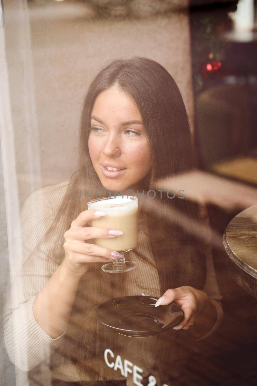 A beautiful girl sits in cafe and looks out window thoughtfully. Reflection of city in window. Brunette woman with long hair drinks cappuccino coffee, vertical, close up