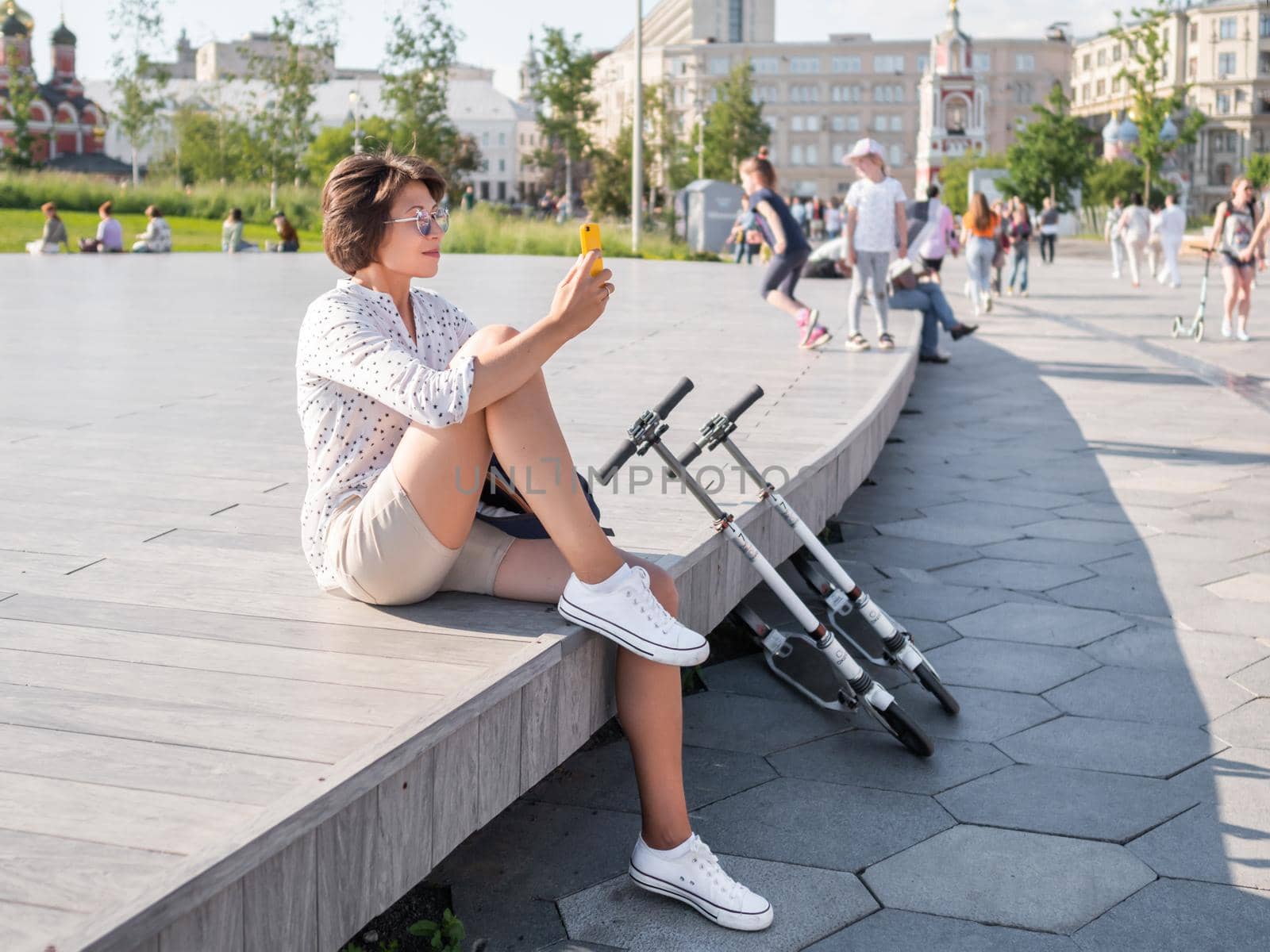 MOSCOW, RUSSIA - June 13, 2021. Woman sits on wooden open stage in public park. Female is making selfie on smartphone after riding kick scooter, eco-friendly urban transport.
