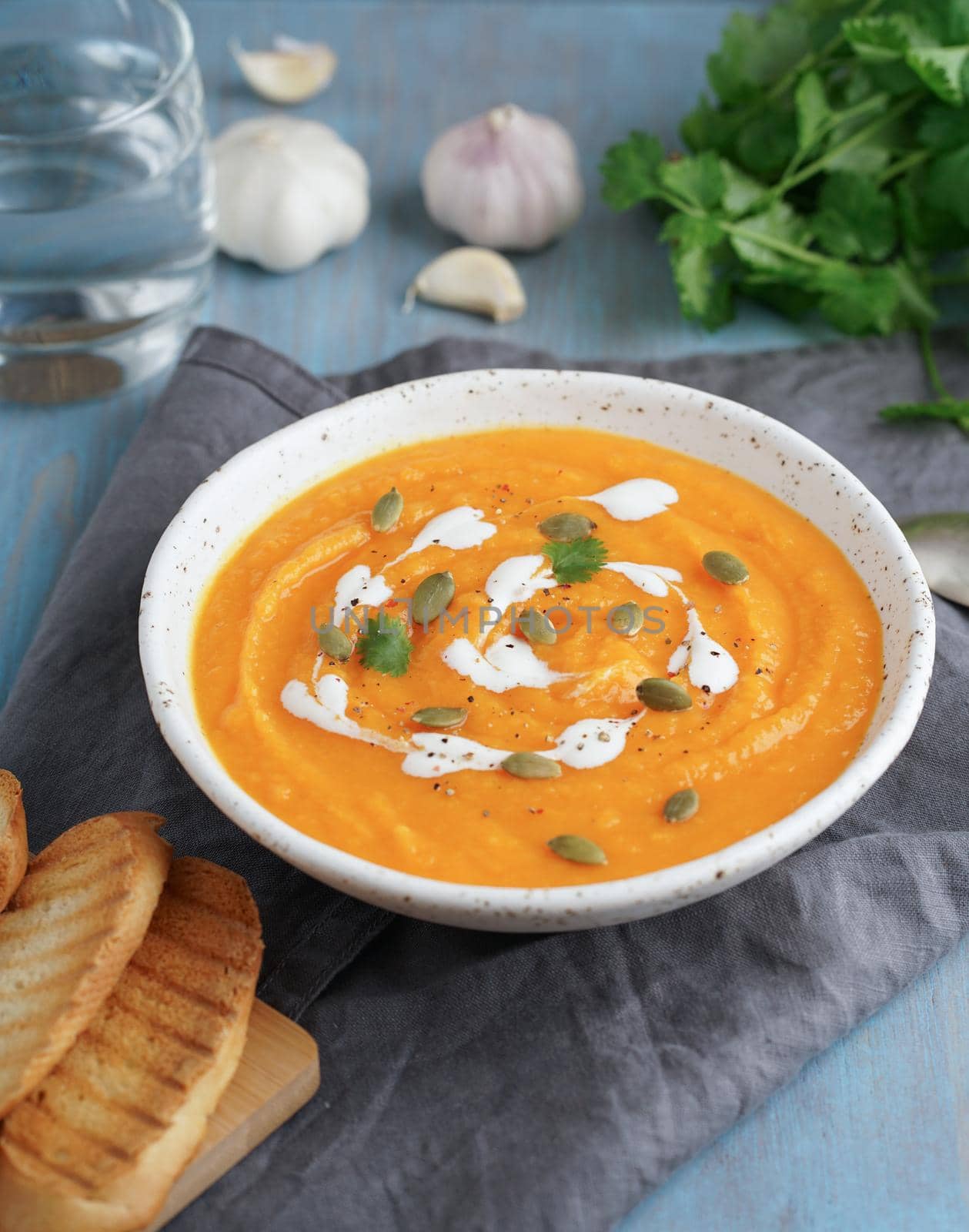 Pupmkin cream soup puree, dietary vegetarian lunch on blue wooden table, vertical. Glass of water, toast, parsley, garlic.