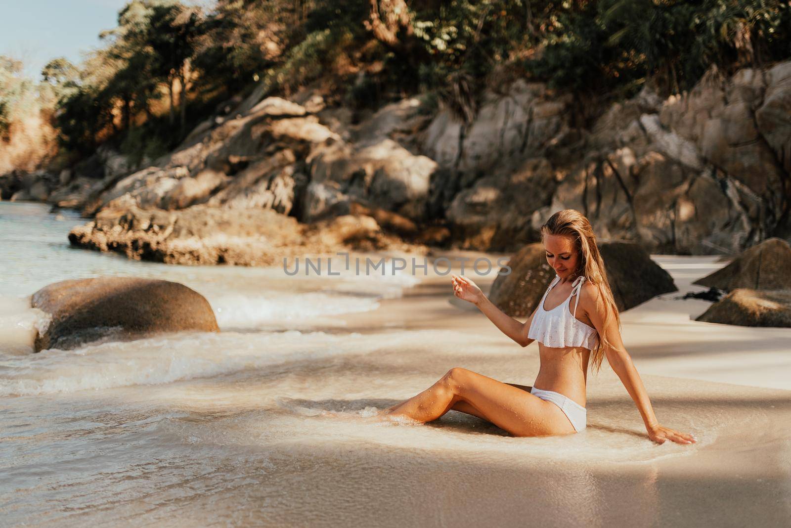 young blond European woman in white bikini swimsuit on beach sitting on sand beach, sea on background of large stone rocks and palm trees