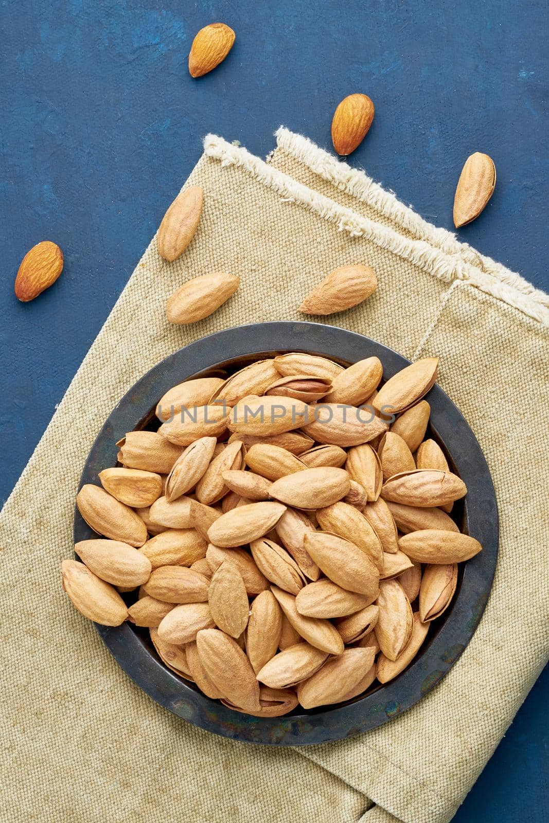 Top view plate with almonds in endocarp, bowl with drupe in shell on a linen napkin on a dark blue background, overhead, copy space, close up, vertical