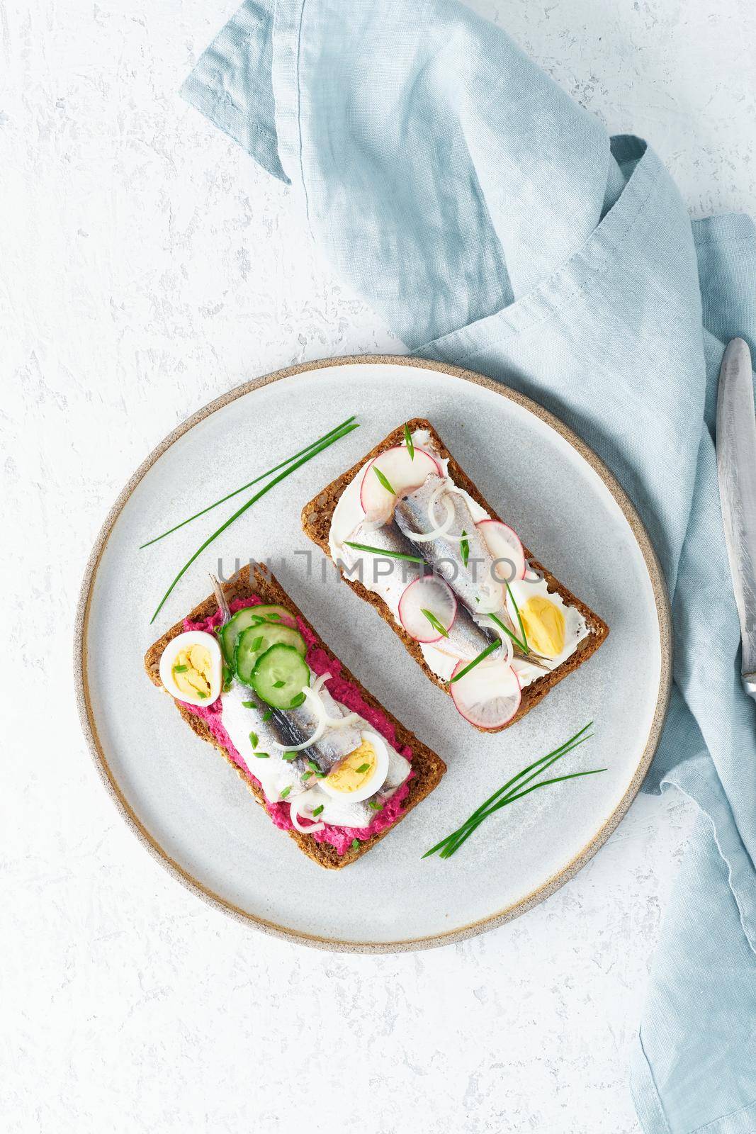 Savory smorrebrod, two traditional Danish sandwiches. Black rye bread with anchovy, beetroot, radish, eggs, cream cheese on grey plate on a white stone table, vertical