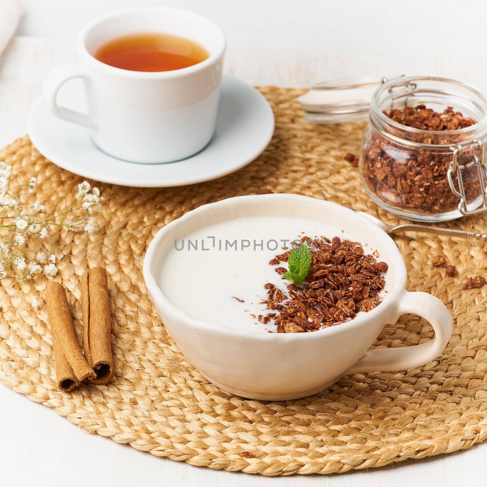 Yogurt with chocolate granola in cup, breakfast with tea on beige background, side view by NataBene