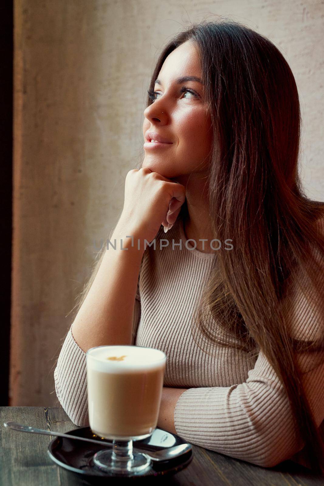 Beautiful pensive happy girl sitting in cafe in Christmas holidays, smiling and dreaming. Brunette woman with long hair drinks cappuccino coffee, latte and looks out window, vertical, dark backdrop