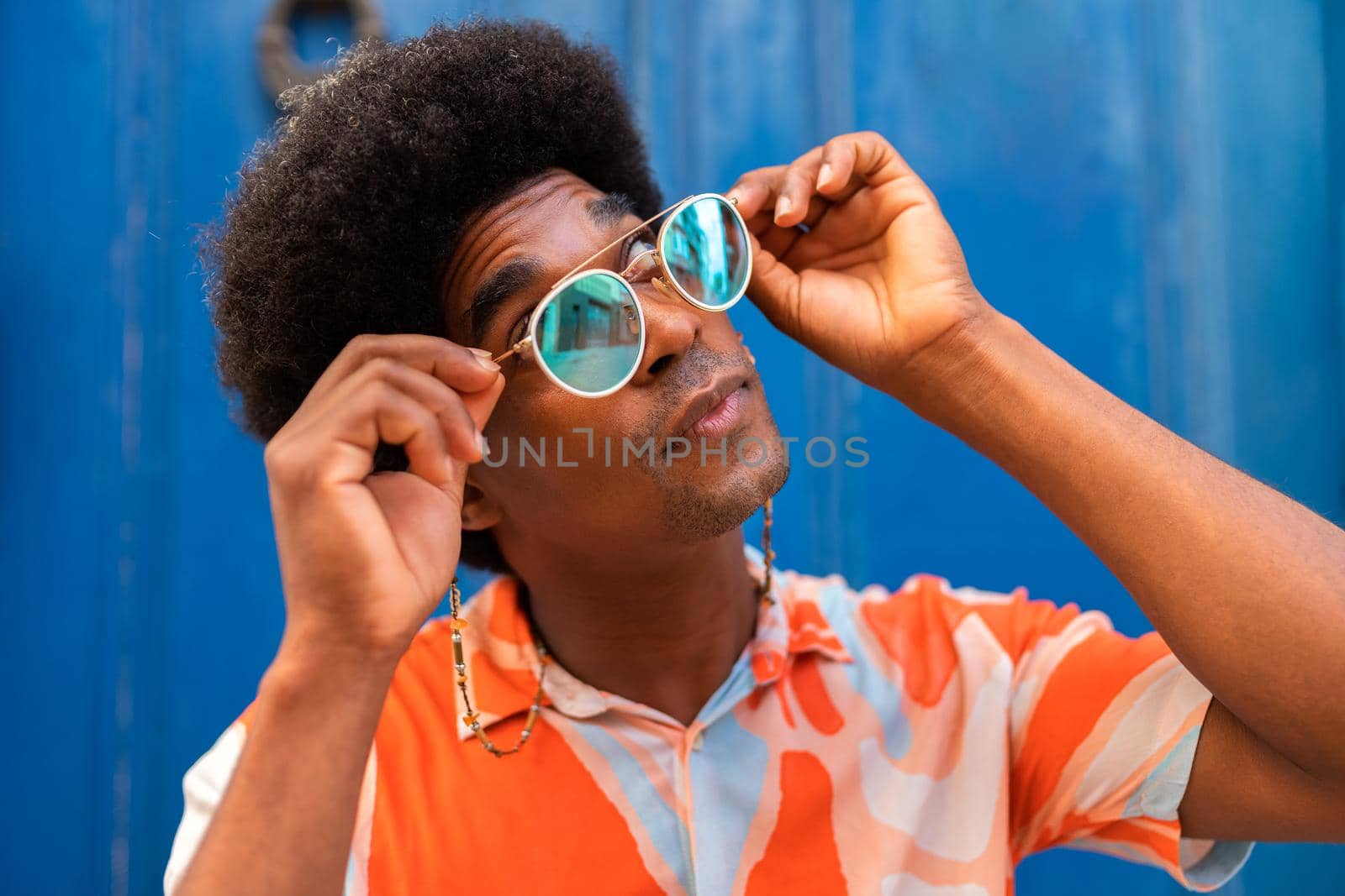 Confident young black man with afro hairstyle putting on sunglasses outdoors. by Hoverstock