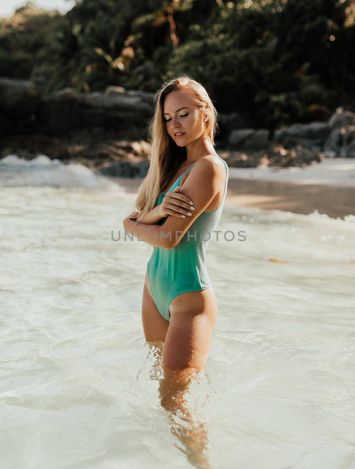 blonde young European woman in bikini swimsuit standing in ocean water, sea on background stone rocks and palms tree