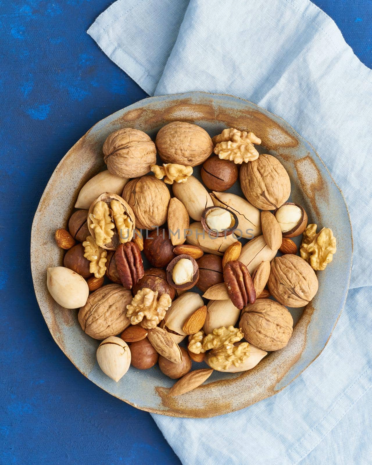 Classic blue in food. Mix of nuts on a plate - walnut, almonds, pecans, macadamia and knife for opening shell. Healthy vegan food. Clean eating, balanced diet. Top view, vertical