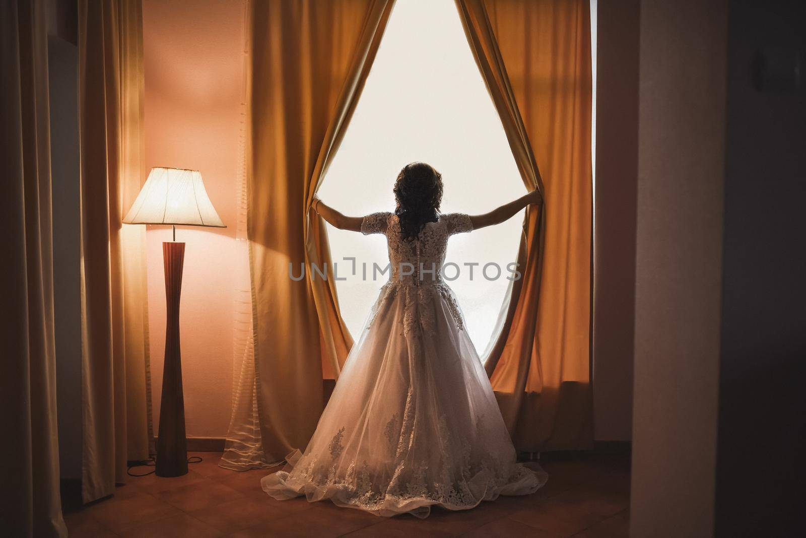 The bride opens the curtains on the huge white window. A dark beige room with low light from a long standing lamp with a shade.