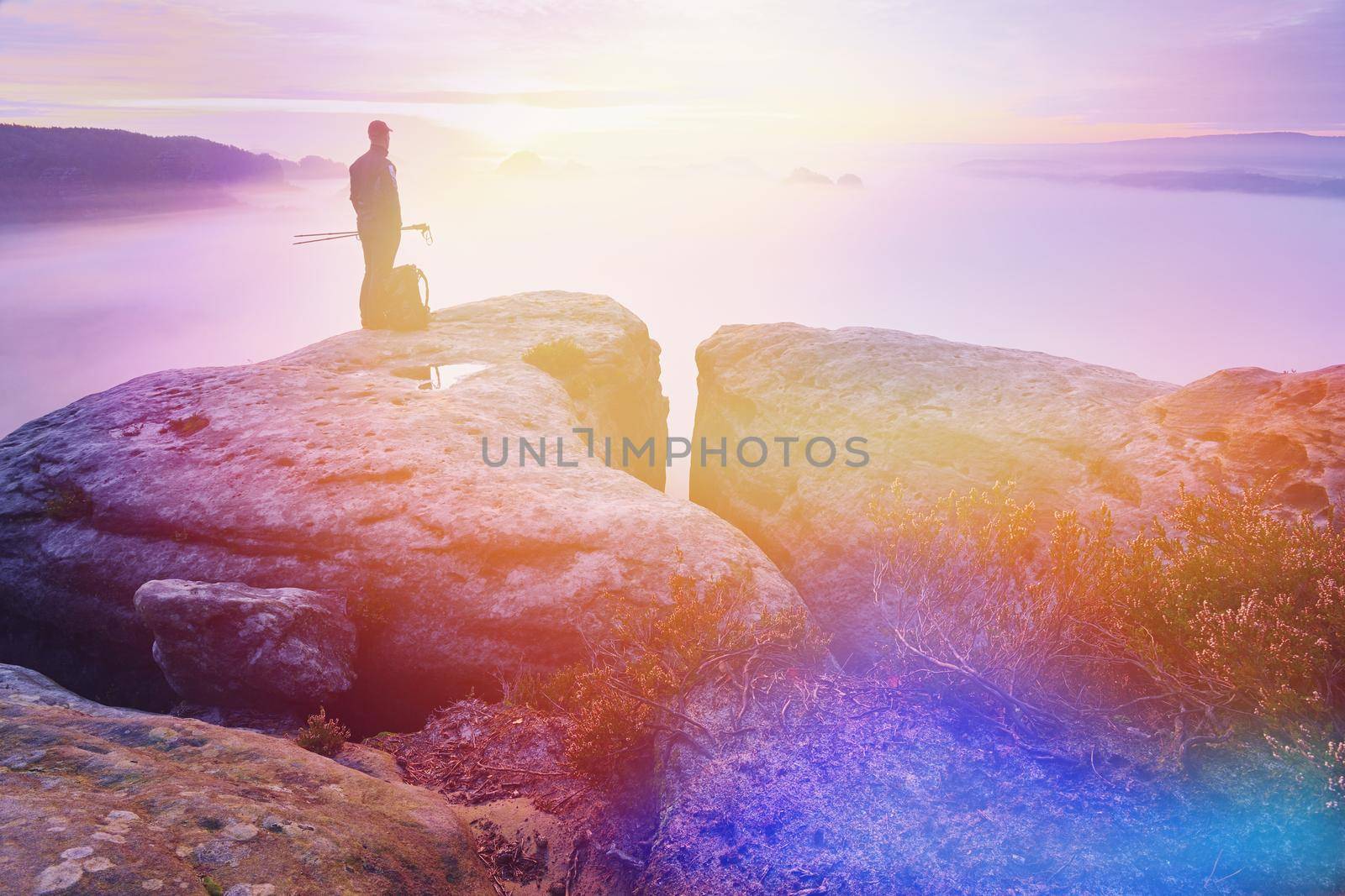 Man with trekking poles on rocky cliff overlooking the misty valley below mountain.  Abstract lighting, colorful flare.