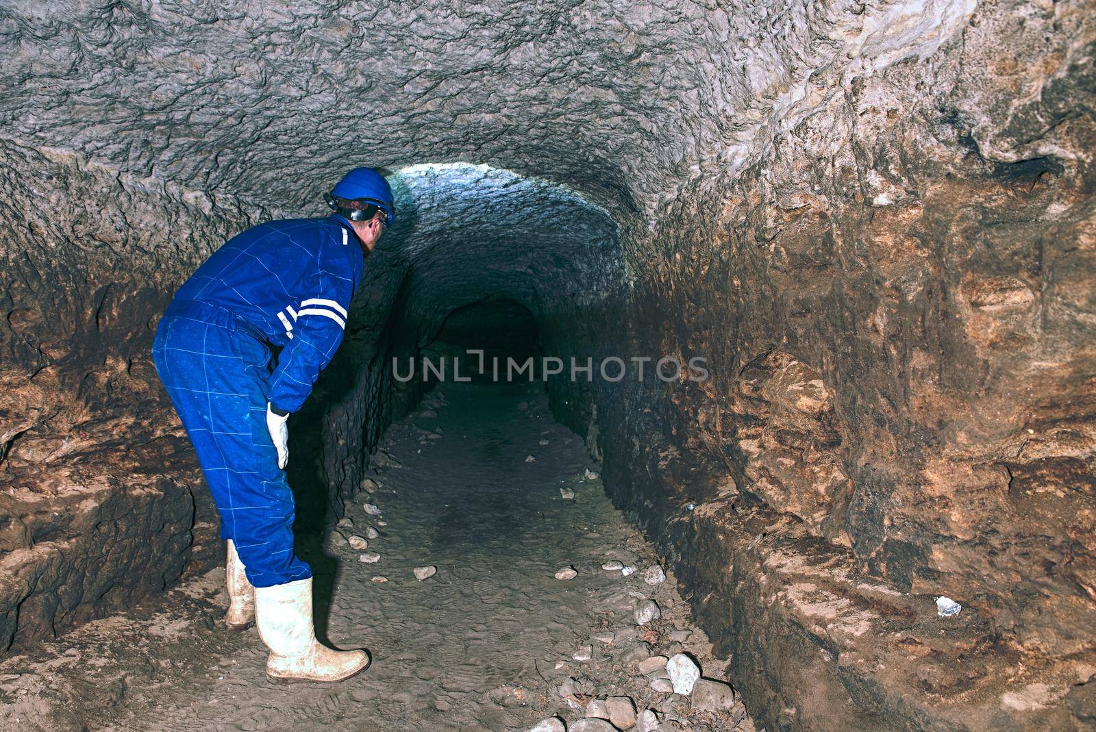 Staff do job in natural underground dome. Man with the illuminated headlight looks into the dark tunnel.