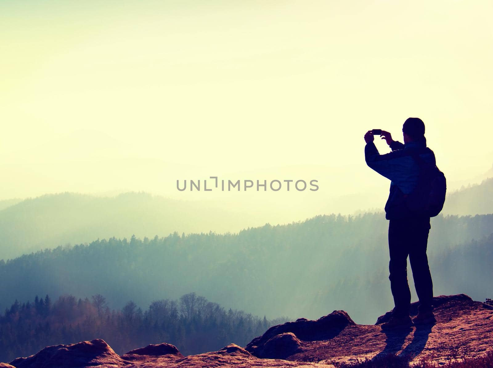 Tourist takes photos with smart phone on peak of rock. Dreamy fogy landscape, spring orange pink misty sunrise in a beautiful valley below rocky mountains. by rdonar2