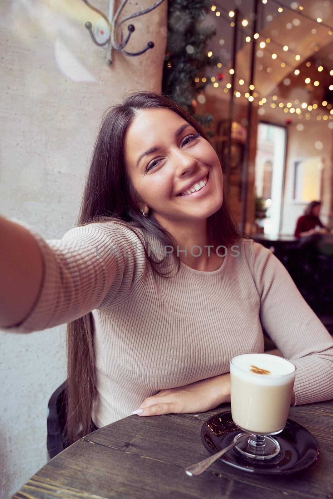 Beautiful happy girl taking a selfie in cafe during Christmas holidays, smiling and looking at phone. Brunette woman with long hair drinks cappuccino coffee, latte, vertical