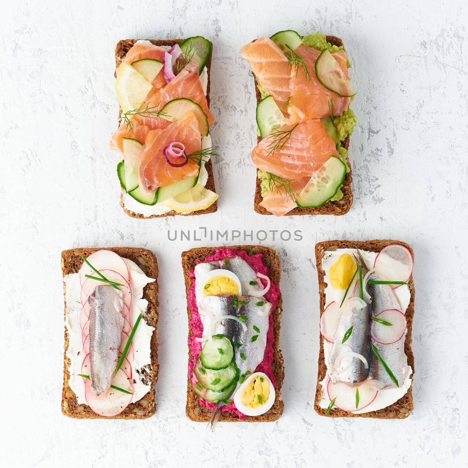 Savory fish smorrebrod, set of five traditional Danish sandwiches. Black rye bread with anchovy, beetroot, radish, eggs, salmon, cream cheese, cucumber, avocado on a grey white stone table, top view