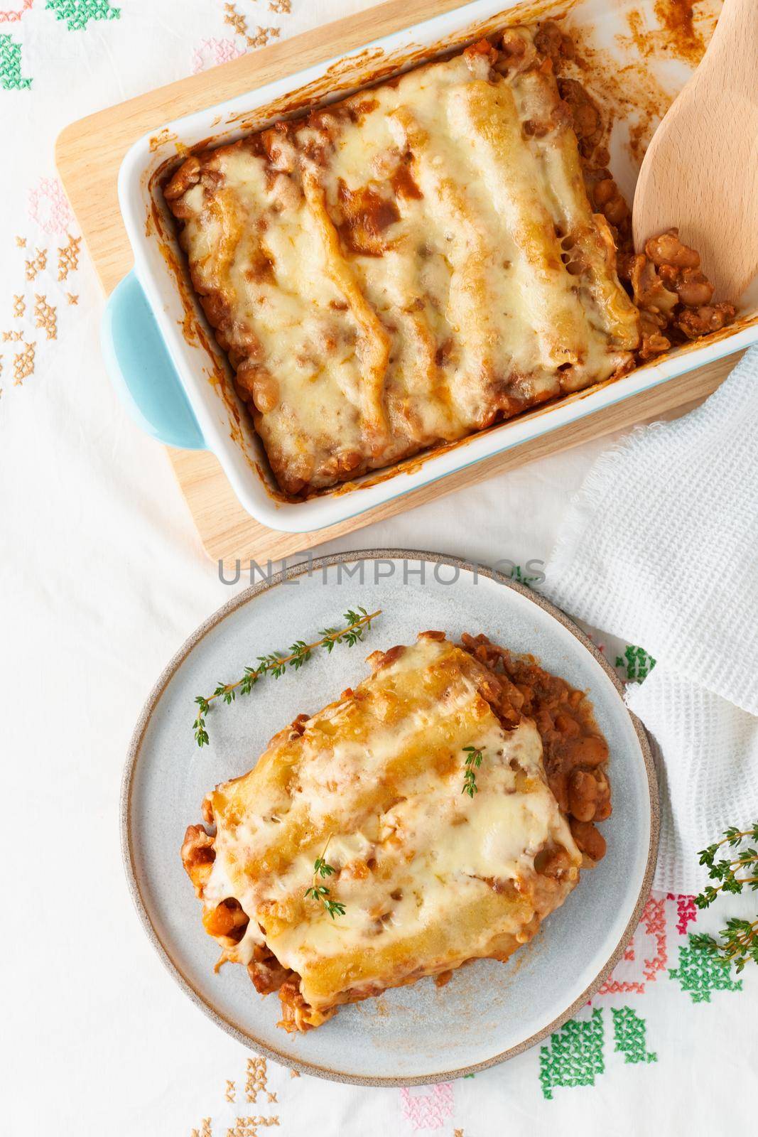 Cannelloni pasta with filling of ground beef, tomatoes, baked with bechamel tomato sauce and a mozzarella. Classical Italian cuisine, white tablecloth with embroidery, rustic style, top view, vertical